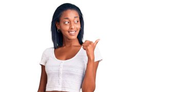 Genç Afro-Amerikan kadın, günlük kıyafetler giyip gülümsüyor, gülümsüyor ve başparmağıyla yan tarafı işaret ediyor.. 