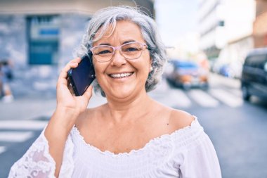 Gri saçlı orta yaşlı bir kadın mutlu bir şekilde dışarıda telefonda konuşuyor.