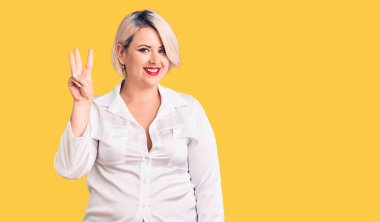 Genç sarışın ve cüsseli kadın rahat gömlekler giyiyor ve üç numaralı parmaklarıyla işaret ederken kendinden emin ve mutlu gülümsüyor.. 