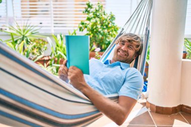 Orta yaşlı yakışıklı bir adam evinin terasında bir hamakta uzanıp kitap okuyor.