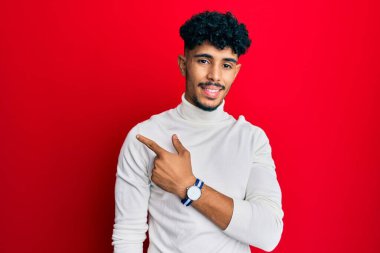 Balıkçı yaka kazak giyen genç Arap yakışıklı adam. Gülümsüyor, eli ve parmağı yana doğru uzanıyor. 