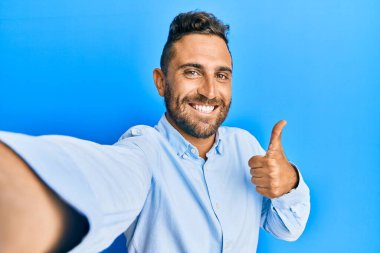 Sakallı yakışıklı bir adam akıllı telefonuyla fotoğraf çekiyor mutlu ve pozitif gülümsüyor, başparmağı mükemmel ve onay işareti yapıyor. 