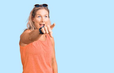 Orta yaşlı, formda, yaz kıyafetleri giyen ve güneş gözlüklü bir kadın. Parmağıyla işaret ediyor. Önünde şaşırmış bir ifade, ağzında şaşkınlık ifadesi var. 