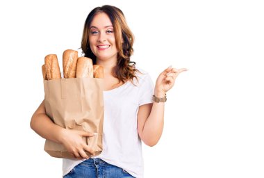 Genç, beyaz, güzel bir kadın, elinde ekmek torbası, gülümsüyor, mutlu bir şekilde elini ve parmağını yana doğru gösteriyor. 