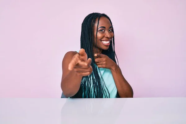 African american woman with braids wearing casual clothes sitting on the table pointing fingers to camera with happy and funny face. good energy and vibes.