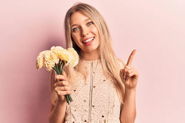 Mladá Blondýna Žena Drží Květiny Úsměvem Šťastný Ukazující Rukou Prstem — Stock fotografie