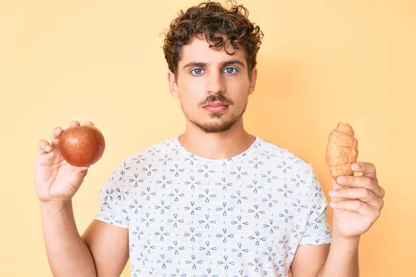 Kıvırcık saçlı, elma ve kruvasan tutan genç beyaz adam yüzünde ciddi bir ifadeyle rahatlamış. Basit ve doğal bir şekilde kameraya bakıyorsun.. 