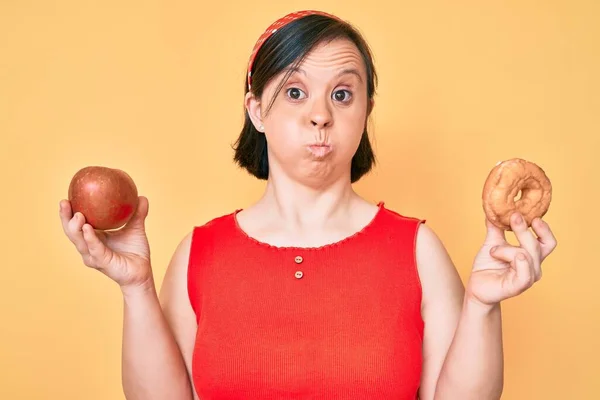 Brunette woman with down syndrome holding red apple and donut sitting puffing cheeks with funny face. mouth inflated with air, catching air.