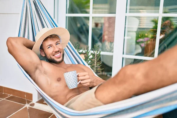 英俊的高加索人高兴地微笑着 躺在阳台上的吊床上 戴着夏帽 喝着一杯咖啡 — 图库照片