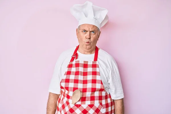Senior grey-haired man wearing professional baker apron making fish face with lips, crazy and comical gesture. funny expression.