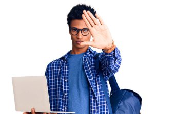 Genç Afro-Amerikalı bir adam sırt çantasını elinde laptopuyla tutuyor ve elinde dur işareti ile ciddi ve kendinden emin bir ifadeyle savunma jesti yapıyor. 