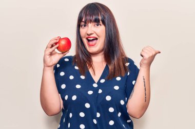 Genç ve cüsseli bir kadın kırmızı elmayı tutup baş parmağını yana uzatıyor ve ağzı açık gülümsüyor. 