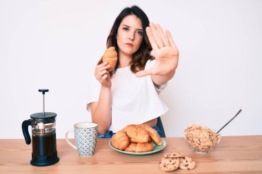 Genç, güzel, esmer bir kadın kahvaltı yapıyor, elinde kruvasanla dur işareti yapıyor, ciddi ve kendinden emin bir ifadeyle, savunma jesti yapıyor. 