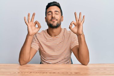 Masaya oturmuş, rahat rahat gülümseyen, gözleri kapalı meditasyon yapan ve parmaklarıyla meditasyon yapan yakışıklı İspanyol adam. yoga konsepti. 