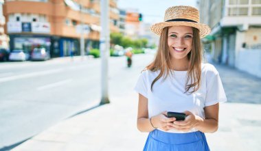 Genç sarışın kadın tatilde gülümsüyor. Şehrin caddesinde akıllı telefon kullanıyor.