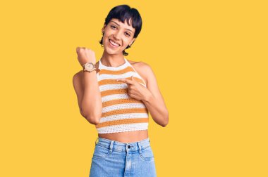 Kısa saçlı genç esmer kadın, günlük kıyafetler giyiyor. Aceleyle saati gösteriyor, sabırsız, rahat bir ifadeyle kameraya bakıyor. 