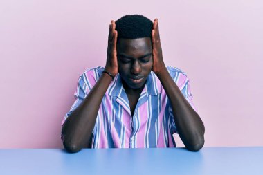 Genç, Afro-Amerikan bir adam, gündelik giysiler giymiş, baş ağrısı yüzünden çaresiz ve stresli bir şekilde masada oturuyor. Çünkü acı ve migren ağrısı var. Eller başının üstünde.. 