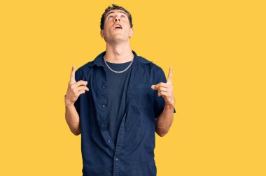 Gündelik giysiler giyen genç yakışıklı adam şaşırmış ve şaşırmış bir şekilde yukarı bakıyor ve parmaklarıyla işaret ediyor ve kollarını kaldırıyor.. 