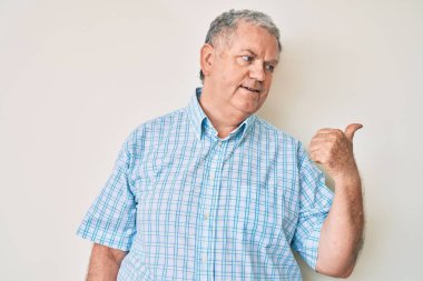 Yaşlı, gri saçlı, günlük kıyafetler giyen, gülümseyen, gülümseyen ve baş parmağıyla yüzünü gösteren bir adam.. 