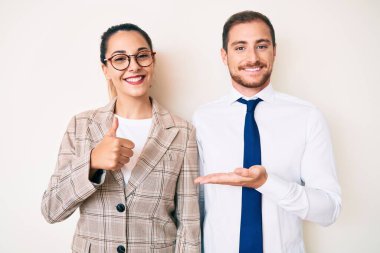 Güzel bir çift iş kıyafetleri giyer, avuç içini gösterir ve baş parmaklarıyla güzel hareketler yapar, mutlu ve neşeli gülümserler. 