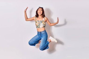 Genç, güzel Çinli kız günlük kıyafetler ve güneş gözlüğü takıp mutlu gülümsüyor. İzole edilmiş beyaz arka planda yüzünde gülümsemeyle zıpla