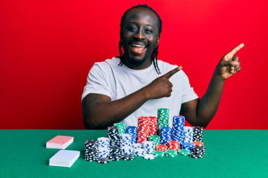 Yakışıklı genç siyah adam poker fişleri ve kartlarla masada oturuyor gülümsüyor ve kameraya bakıyor iki eliyle ve parmaklarıyla kenarı işaret ediyor.. 