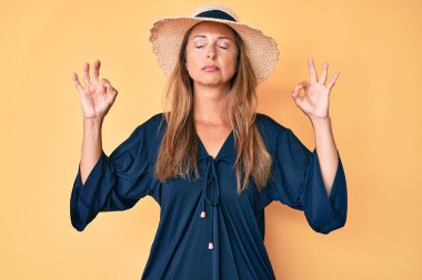 Orta yaşlı İspanyol bir kadın yaz şapkası takıyor. Gözleri kapalı gülümsüyor. Parmaklarıyla meditasyon hareketi yapıyor. yoga konsepti. 