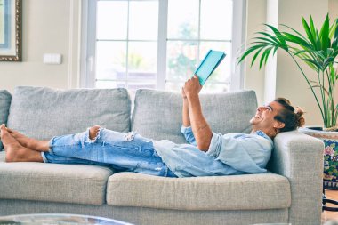 Genç İspanyol gülümseyen mutlu bir kitap okuyucu evdeki kanepede uzanıyor.