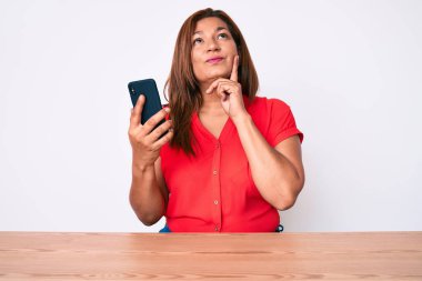 Orta yaşlı esmer bir kadın akıllı telefon kullanıyor. Masada oturuyor. Ciddi bir yüz ifadesi var. 