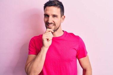 İzole edilmiş pembe arka planda duran rahat tişört giyen genç ve yakışıklı bir adam kameraya kollarını kavuşturup çenesine dokunarak gülümsüyor. Olumlu düşünüyorum..