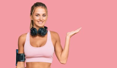 Spor kıyafetleri giyen, kulaklık takan neşeli bir şekilde gülümseyen ve avuç içi kameraya bakan genç sarışın kadın.. 