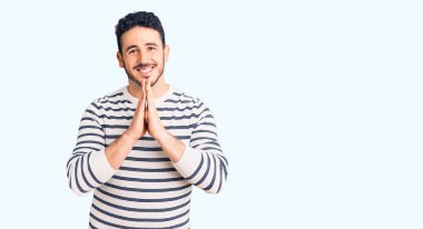 Gündelik kıyafetler giyen genç İspanyol adam elleriyle dua ediyor af diliyor kendinden emin bir şekilde gülümsüyor.. 