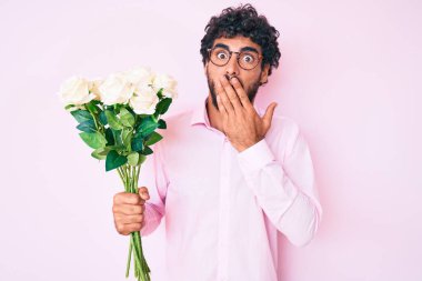 Kıvırcık saçlı yakışıklı genç adam ve elinde bir buket beyaz çiçekle ağzını eliyle kapatıyor, şokta ve hata yapmaktan korkuyor. şaşırmış ifade 