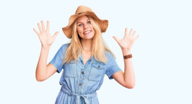 Genç, sarışın, yaz şapkası ve elbisesi takan on numaralı parmaklarıyla onu gösterirken kendine güvenen ve mutlu bir şekilde gülümseyen güzel bir kadın.. 