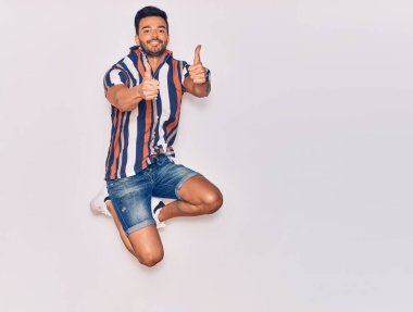 Genç, yakışıklı, sıradan kıyafetler giyen, mutlu gülümseyen bir İspanyol. Yüzünde tebessümle zıpla, tamam işareti yap, başparmağınla izole beyaz arkaplan üzerinde.