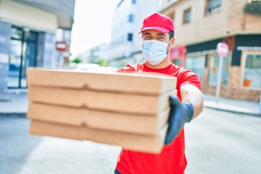Üniforma giyen ve koronavirüs koruma maskesi takan genç teslimatçı. Kasaba caddesinde pizza kutularını teslim ediyorum..