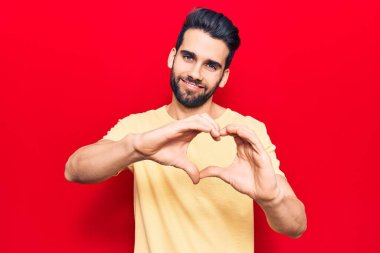 Sakallı, sıradan tişörtlü, aşık bir şekilde gülümseyen, kalp sembolü ve elleri olan genç ve yakışıklı bir adam. romantik kavram. 