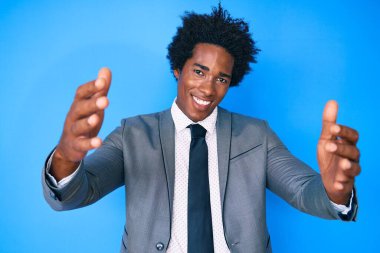 Afro saçlı, iş ceketi giyen yakışıklı Afro-Amerikan adam kameraya bakıp sarılmak için gülümsüyor. Mutluluğu kucaklayan neşeli bir ifade.. 