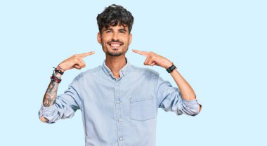 Günlük kıyafetler giyen, neşeli gülümseyen, parmaklarıyla ve ağzıyla işaret eden İspanyol genç bir adam. diş sağlığı kavramı. 