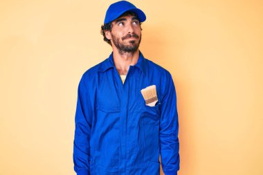 Kıvırcık saçlı ve ayı kılıklı yakışıklı bir genç adam inşaatçı tulumu giyiyor. Gülümsüyor ve yan tarafa bakıyor ve düşünüyor.. 