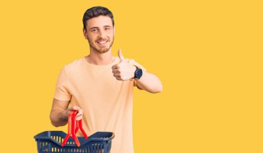 Süpermarket alışveriş sepeti taşıyan yakışıklı genç adam mutlu ve pozitif gülümsüyor, başparmağıyla mükemmel ve onay işareti yapıyor. 