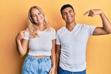 Irklar arası genç bir çift, günlük beyaz tişört giyiyorlar, kendilerine güveniyorlar, gülümsüyorlar, parmaklarını gururlu ve mutlu bir şekilde işaret ediyorlar.. 