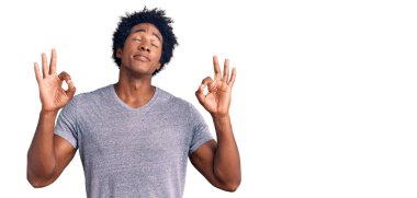 Afro saçlı, rahat giyinen ve gözleri kapalı gülümseyen yakışıklı Afro-Amerikan erkeği parmaklarıyla meditasyon hareketi yapıyor. yoga konsepti. 