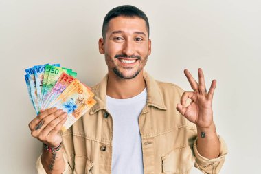 Dövmeleri olan yakışıklı bir adam İsviçre frangı banknotları tutuyor parmaklarıyla imza atıyor, gülümsüyor ve mükemmel bir sembol oluşturuyor. 