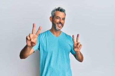 Orta yaşlı, gri saçlı, sıradan kıyafetler giyen, gülümseyen, parmaklarını zafer işareti yaparken gösteren bir adam. İki numara.. 