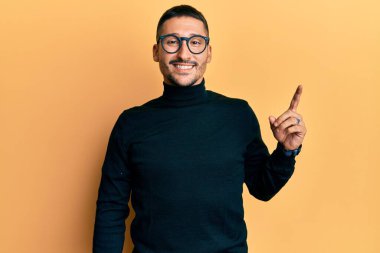 Balıkçı yaka kazak giyen ve yüzünde kocaman bir gülümseme olan gözlüklü yakışıklı bir adam, kameraya el parmağı ile bakıyor.. 