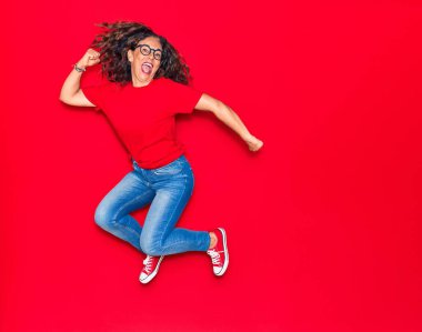 Orta yaşlı, güzel İspanyol bir kadın günlük kıyafetler ve mutlu bir gülümseme takınıyor. İzole edilmiş kırmızı arka planda yüzünde gülümsemeyle zıplıyor