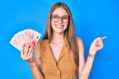 Sarışın genç kız İsrail şekellerini tutuyor gülümsüyor mutlu bir şekilde eli ve parmağı yana doğru gösteriyor 