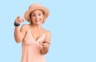 Yazlık şapka takan genç sarışın kadın seni ve kamerayı parmaklarıyla gösteriyor, gülümsüyor ve neşeli. 