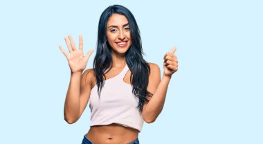 Güzel İspanyol kadın gündelik giysiler giyer ve altı numaralı parmaklarıyla işaret ederken kendinden emin ve mutlu bir şekilde gülümser.. 